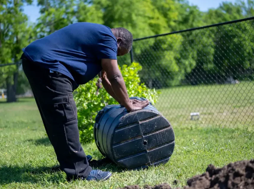 Best compost tumbler — person bending to roll a black barrel-shaped composter across a grass lawn