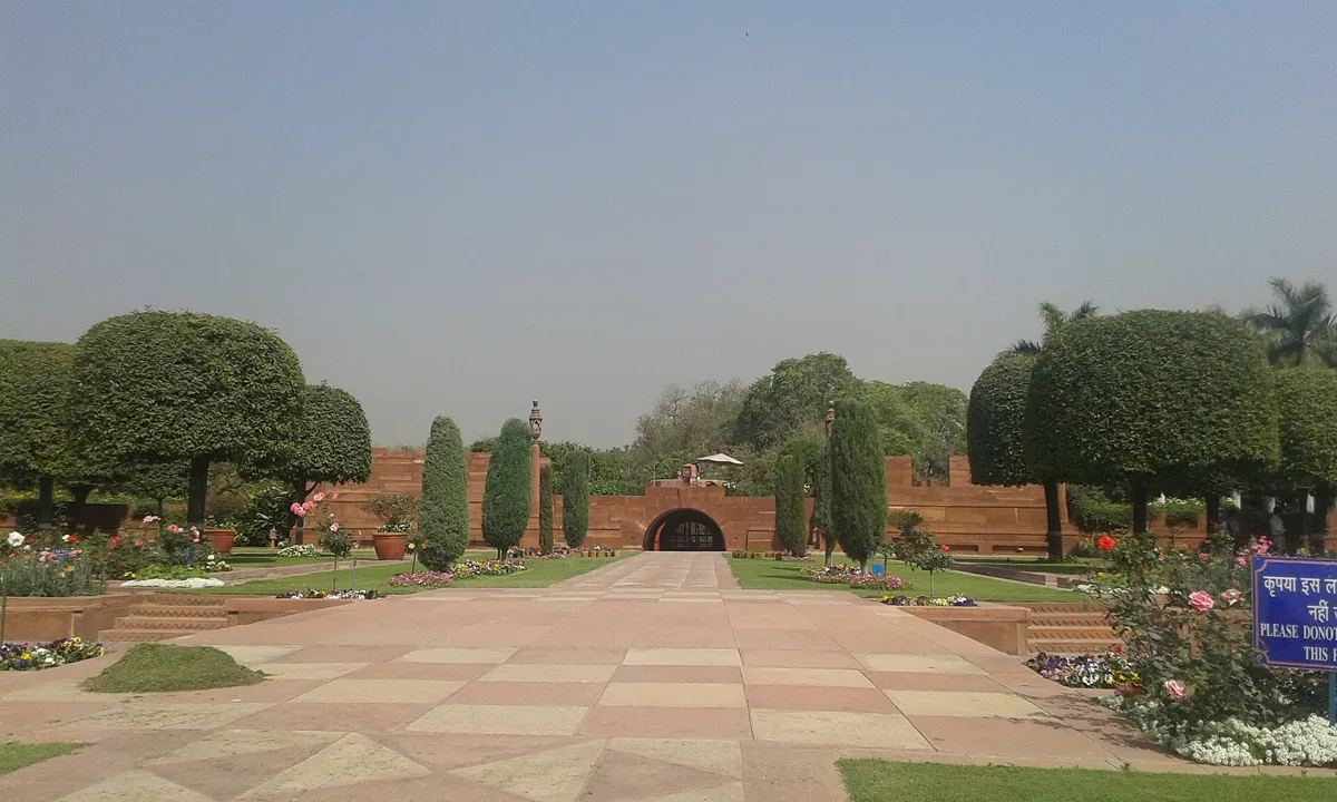 A wide sandstone walkway running between clipped dome-shaped trees and cypresses towards an arched gateway in a red wall