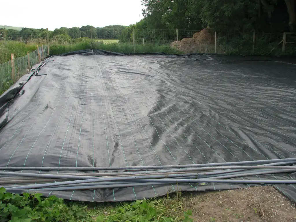 Black woven weed control fabric with green marker lines stretched over a fenced garden plot, with hoops stacked on top ready for use