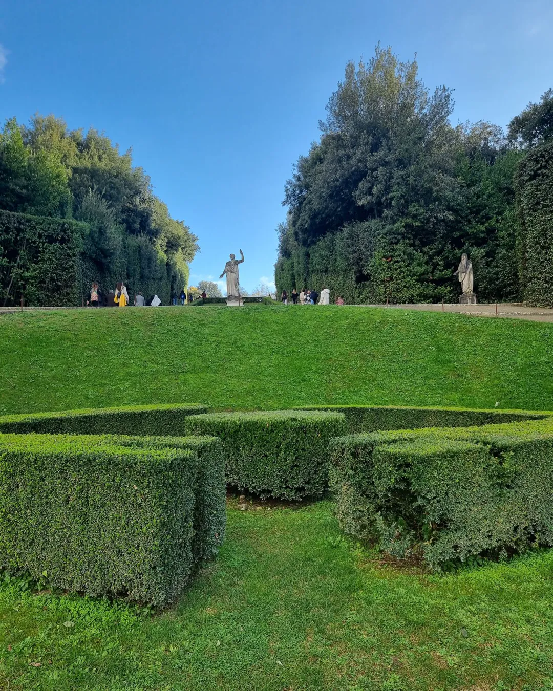 Low clipped box parterre in front of a rising grass bank topped by a statue and hedge wall