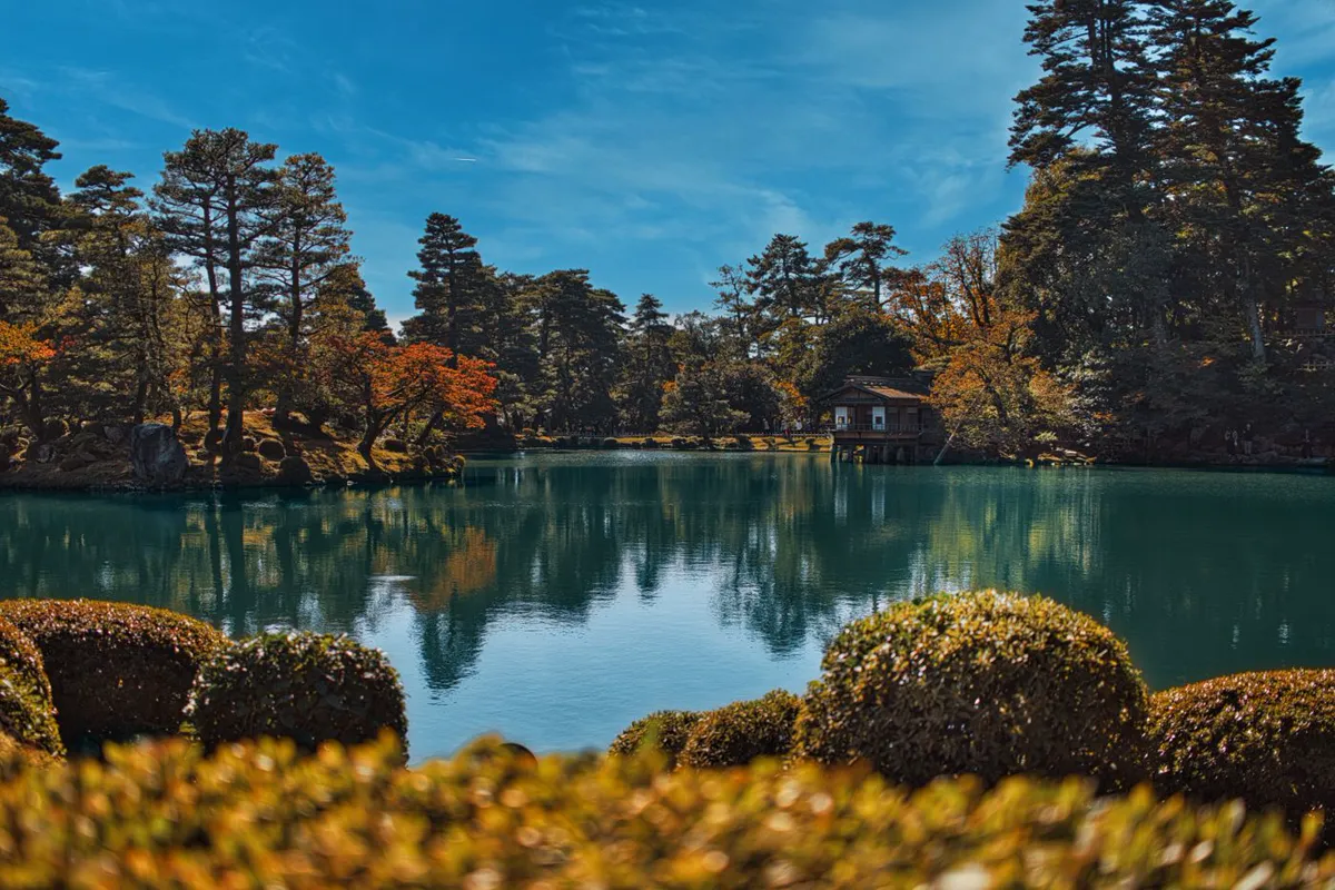 A wide pond reflecting autumn maples, pines and a small wooden teahouse on stilts at the far bank