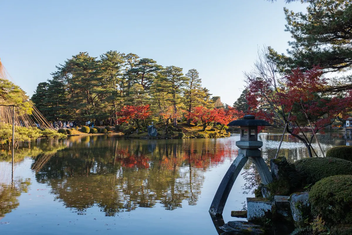 A two-legged stone lantern on a pond bank with red maples, a wooded island and rope snow cones behind